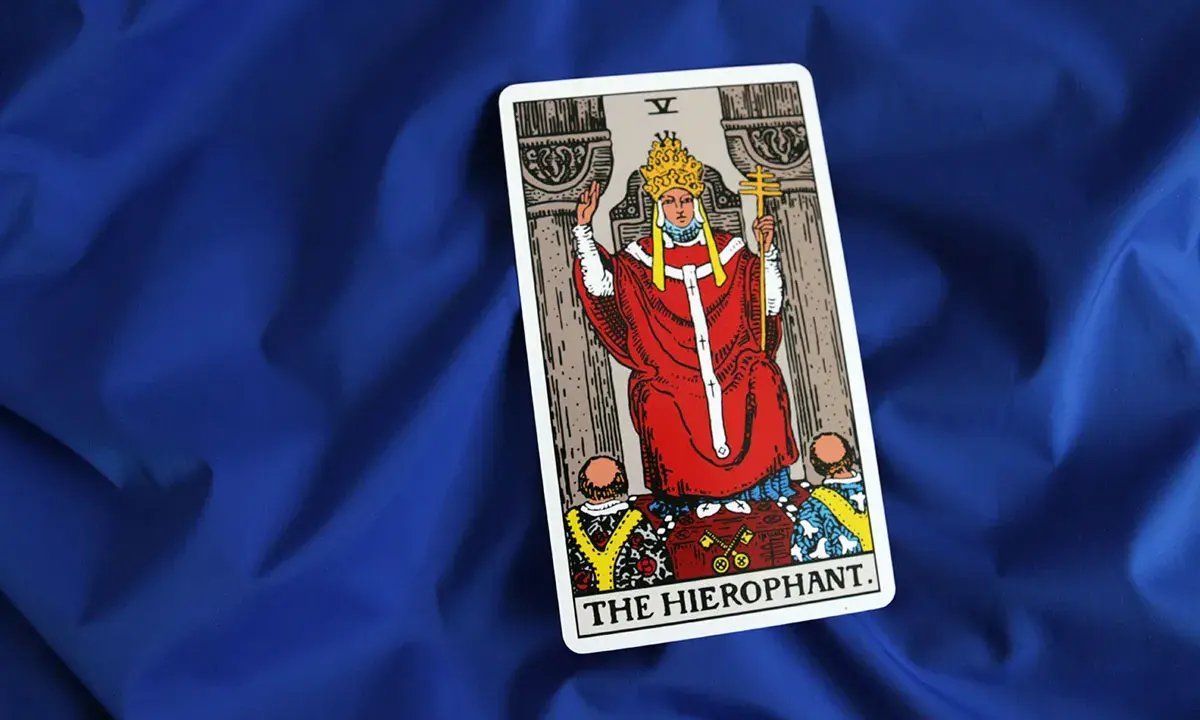Karta Tarota "The Hierophant" na niebieskim tle. Postać w czerwonych szatach z tiarą i krzyżem.