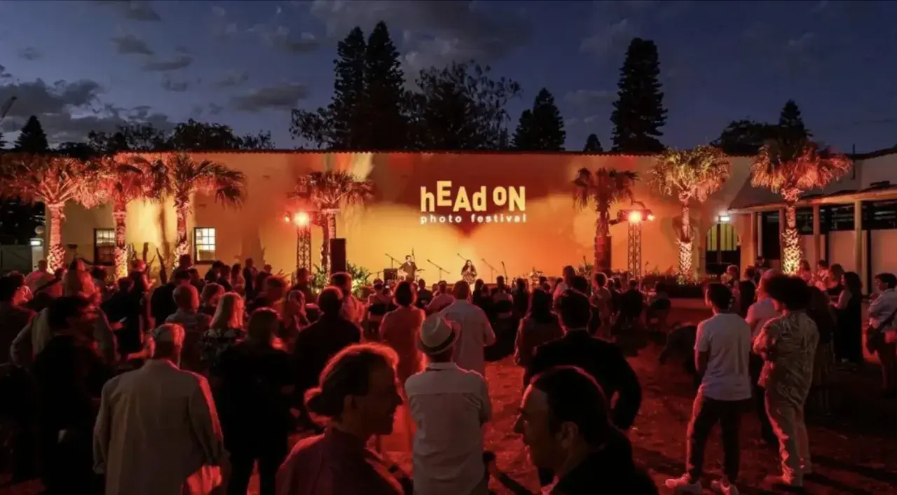 Crowd gathered at the hEAd oN photo festival, enjoying a performance under palm trees lit by warm orange lights.