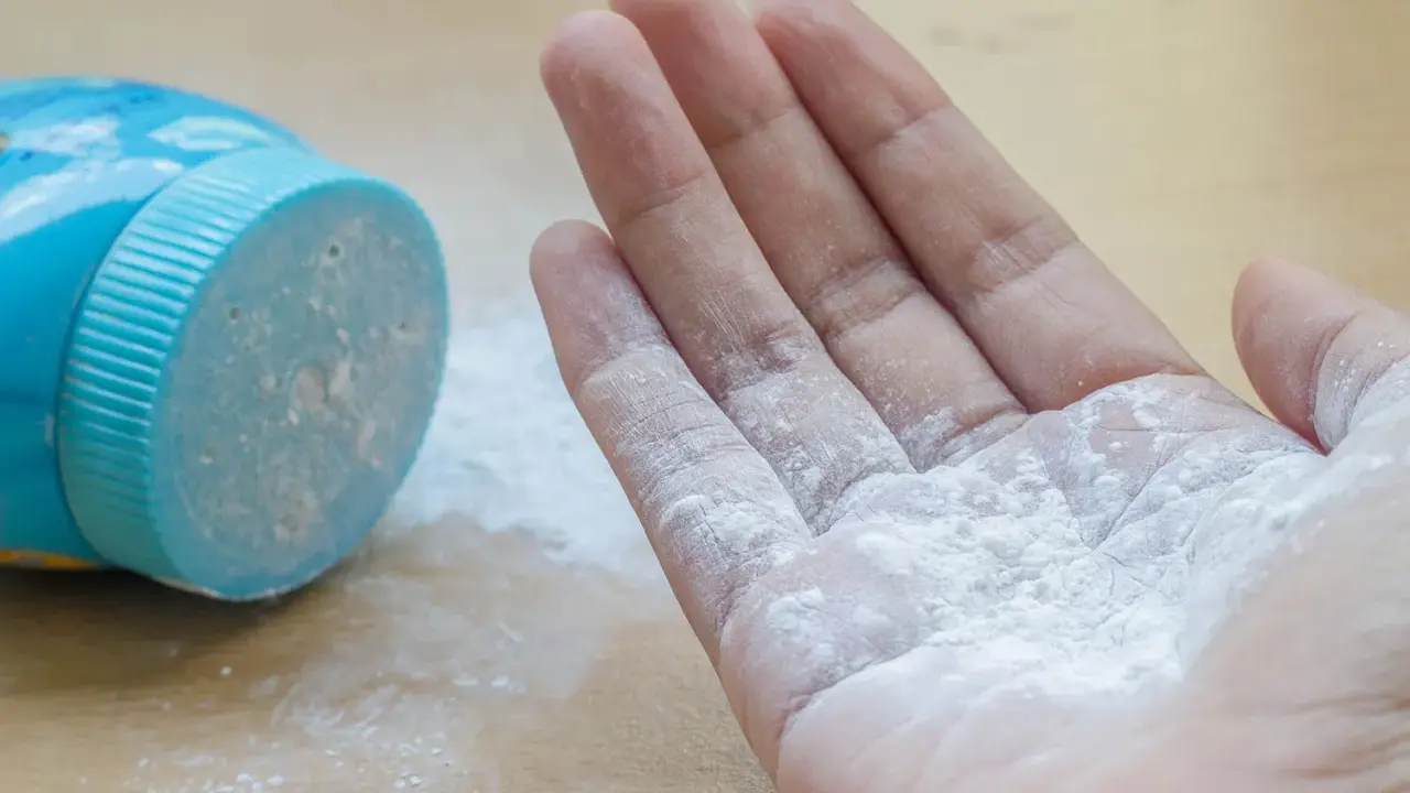 Mano con borotalco bianco sparso, vicino al contenitore blu. Attenzione: il borotalco è cancerogeno.
