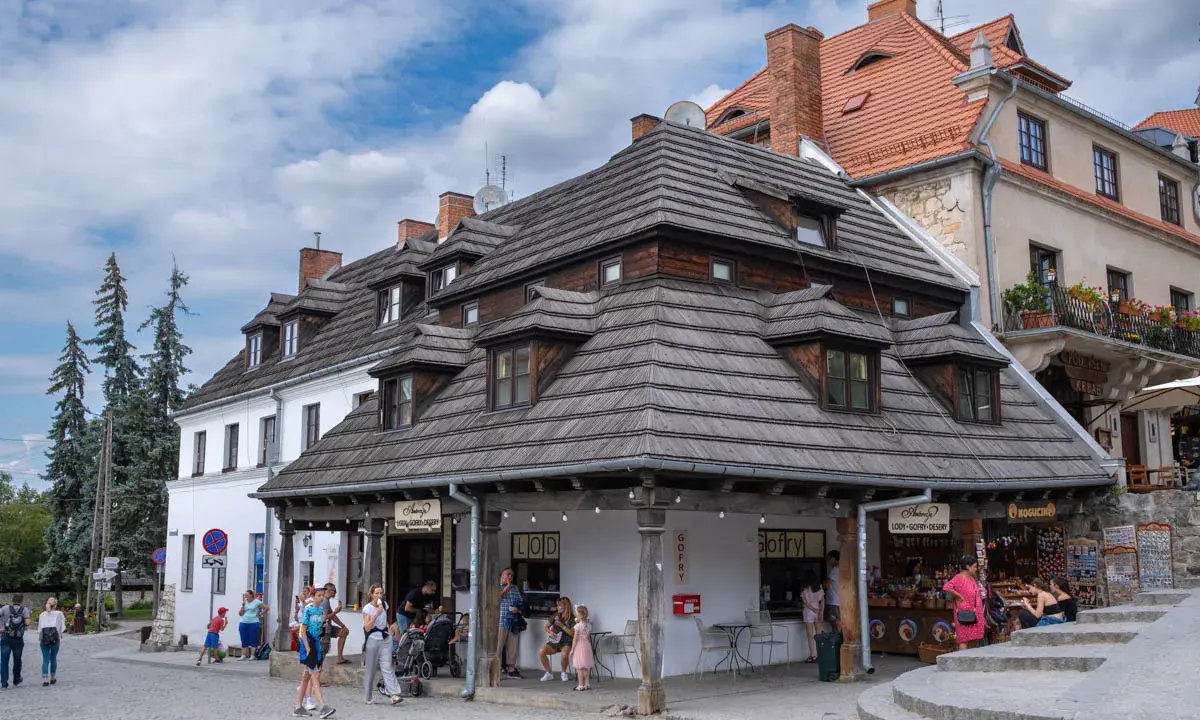 Urokliwy rynek w Kazimierzu Dolnym z charakterystycznymi kamieniczkami i drewnianymi dachami. Ludzie spacerują, kupują lody i pamiątki.