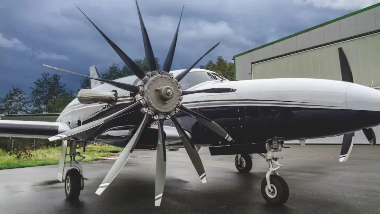 Ein elegantes Flugzeug mit einem beeindruckenden 4-Blatt-Propeller steht auf nassem Asphalt vor einer Halle.