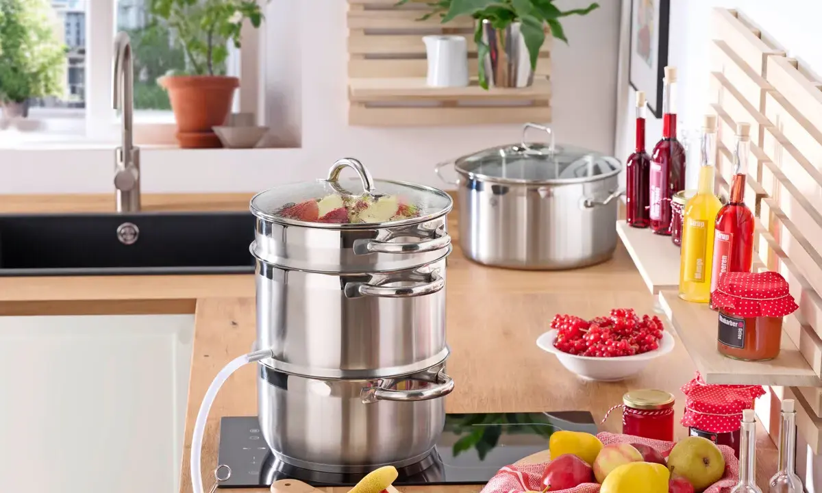 Saft einkochen im Einkochautomat: Obst wird im Dampfgarer zu Saft verarbeitet. Daneben stehen Gläser mit roter Johannisbeermarmelade und Flaschen mit Sirup.