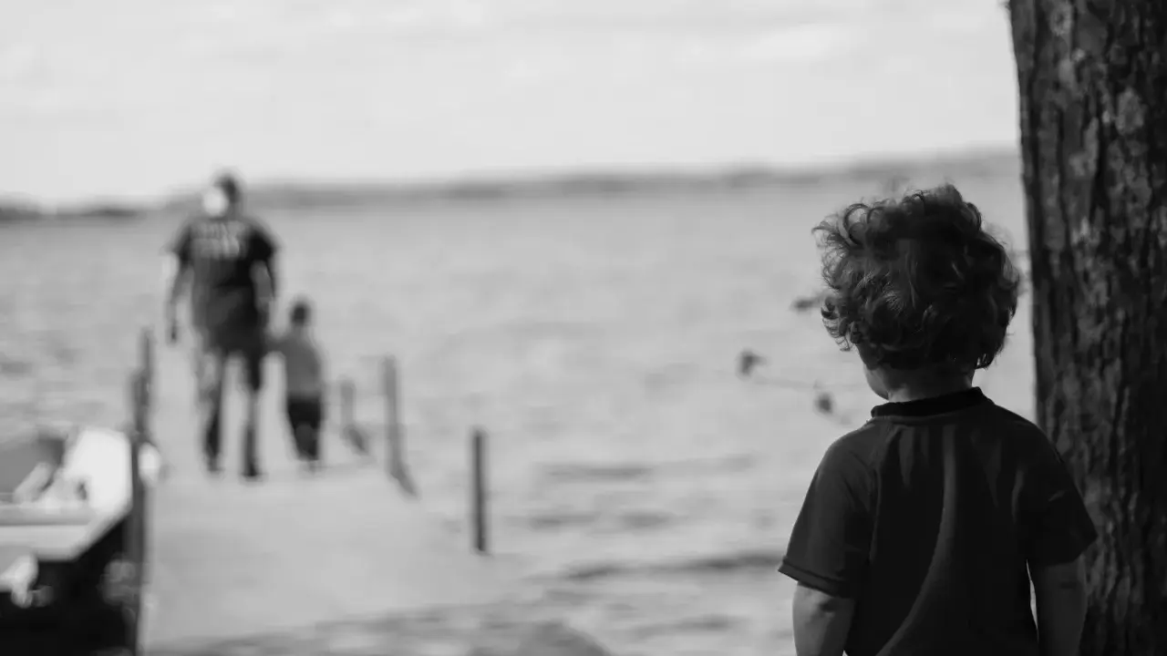 Bambino con capelli ricci guarda verso un molo sfocato dove camminano due figure. Un senso di isolamento, quasi un attaccamento disorganizzato, aleggia nell'aria.