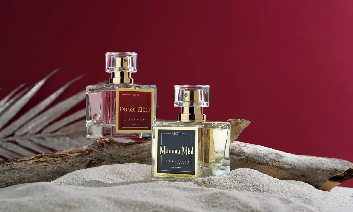 Dwa flakony perfum z etykietami "Dubai Elixir" i "Mamma Mia!", zawierające feromony damskie, stoją na piasku obok kawałka drewna.