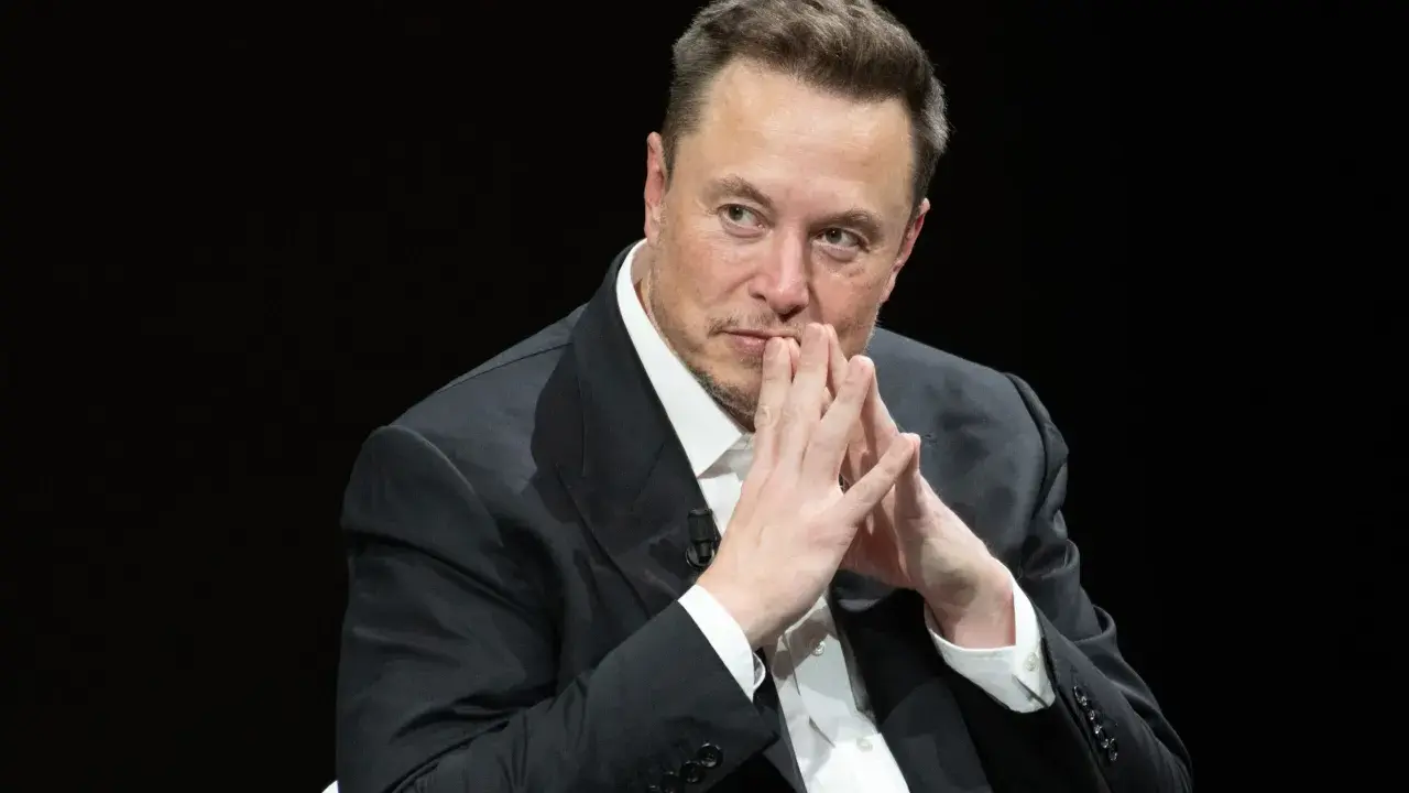 Elon Musk, znany wizjoner i przedsiębiorca, w czarnym garniturze, z dłońmi złożonymi przed twarzą.