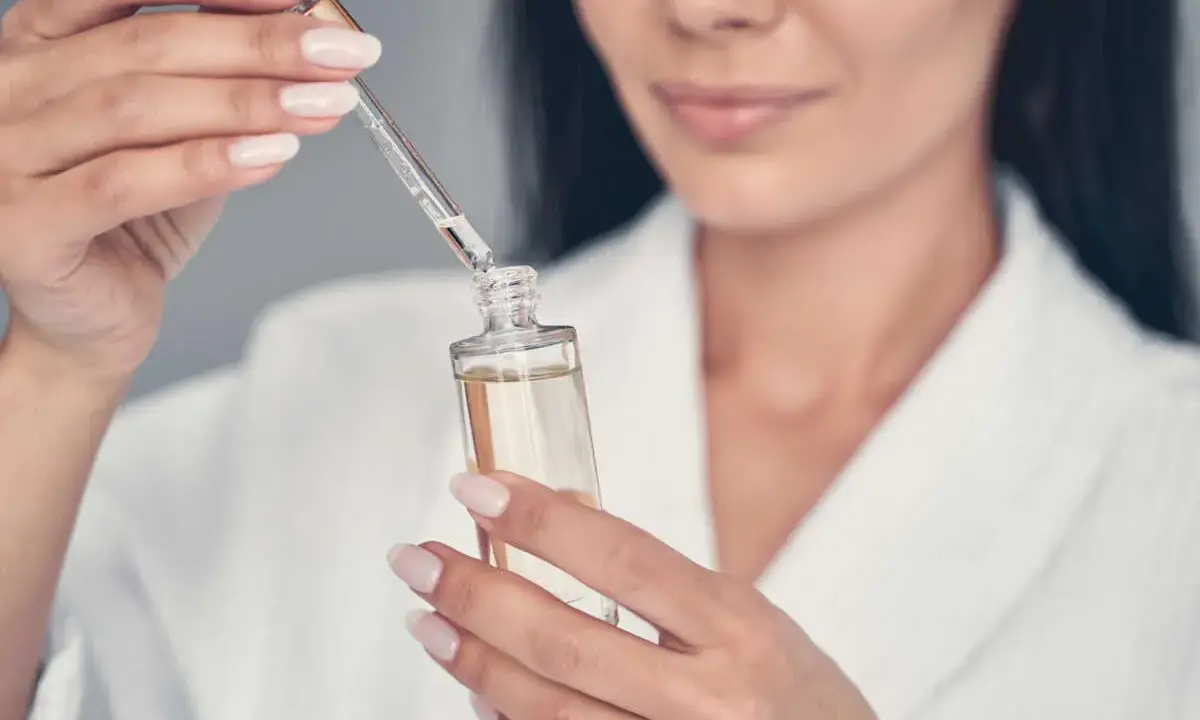 Une femme en peignoir blanc utilise un compte-gouttes pour prélever un sérum, peut-être un mélange de niacinamide et rétinol, pour sa routine de soins.