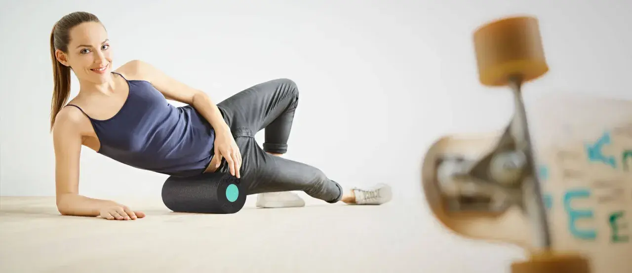 Frau rollt mit einer Faszienrolle über ihr Knie, um Muskelverspannungen zu lösen. Ein Skateboard ist im Vordergrund unscharf zu sehen.