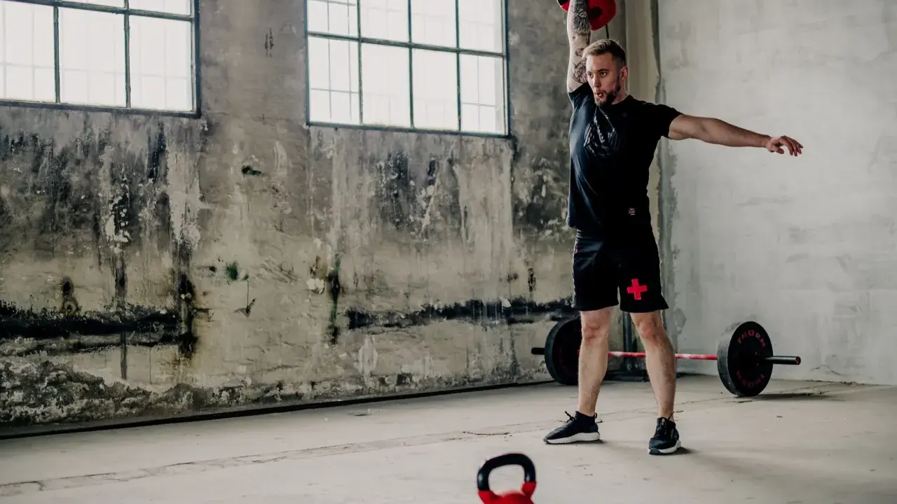 Mężczyzna wykonuje ćwiczenia z kettlebell, idealne dla początkujących. W tle sztanga i surowe wnętrze.