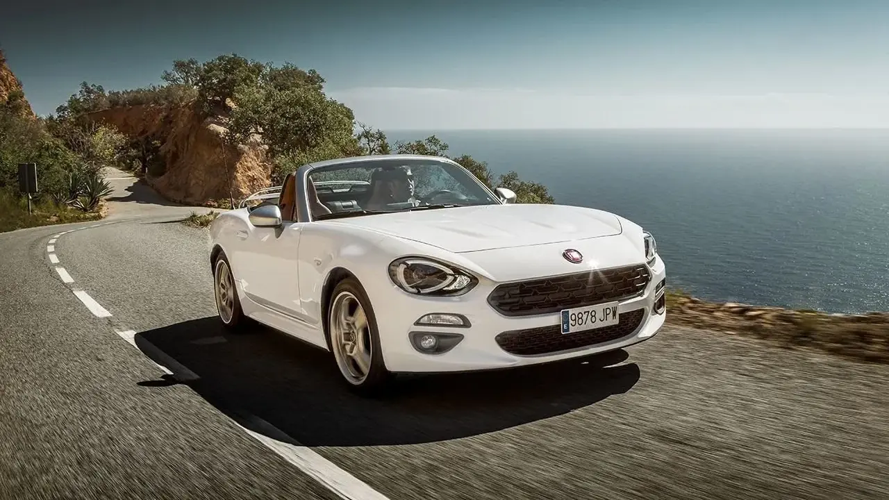 Biały Fiat 124 Spider sunie po krętej drodze nad morzem.