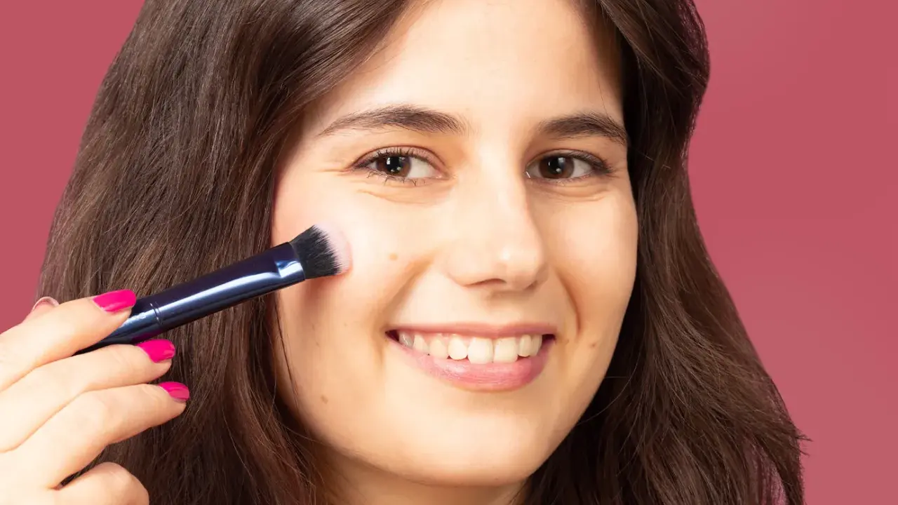 Una mujer sonríe mientras aprende como aplicar colorete con brocha, logrando un rubor natural en sus mejillas.