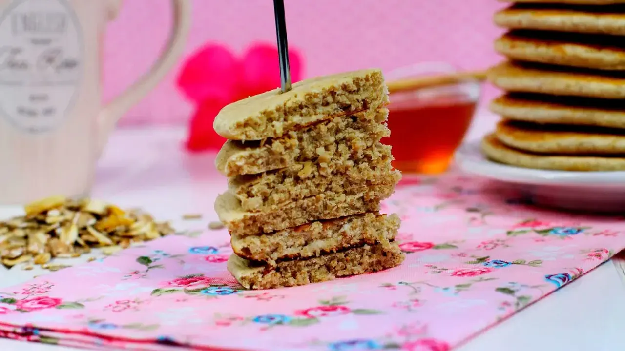 Una pila de tortitas de avena, con un tenedor atravesado, sobre una servilleta floral rosa. Al fondo, una taza y miel.
