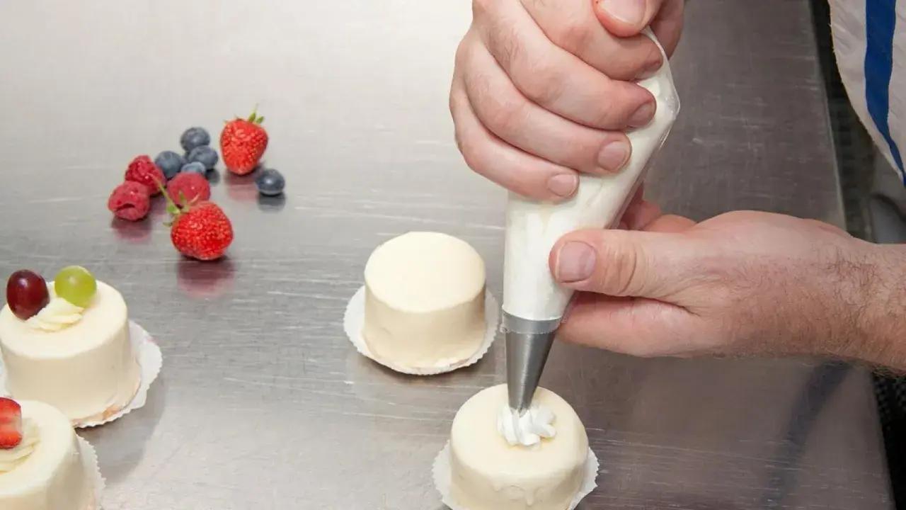 Man usa una manga pastelera para decorar pequeños pasteles. Frutas rojas y arándanos adornan la mesa.