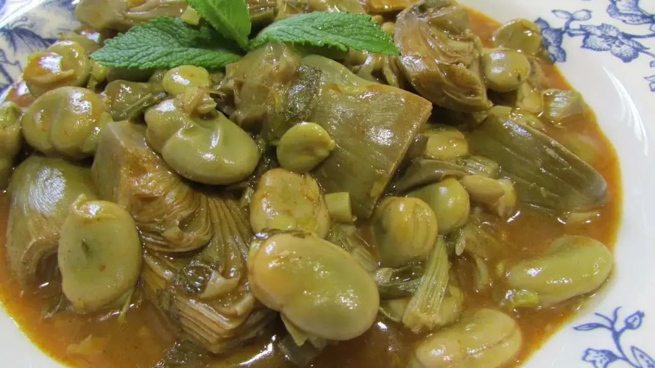 Plato de habas tiernas y trozos de alcachofa en una salsa dorada, decorado con hojas de menta fresca.