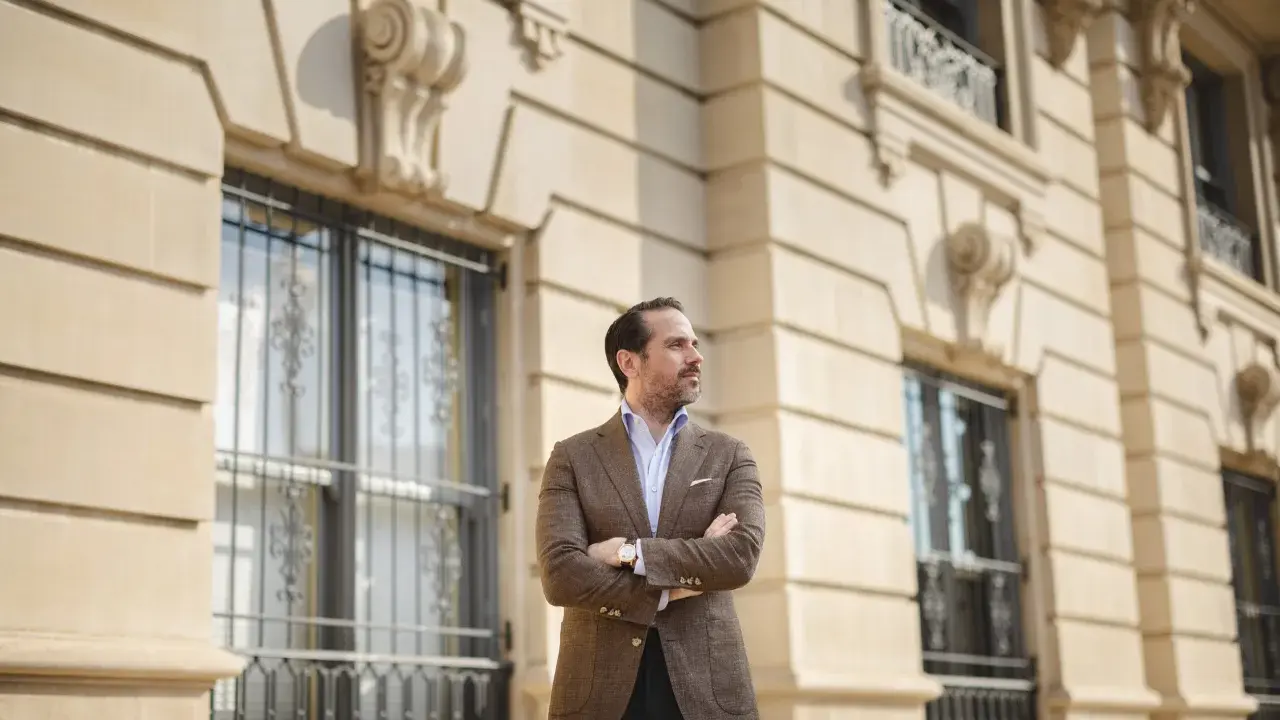 A man in a stylish brown sport coat outfit stands with arms crossed in front of a classic building.
