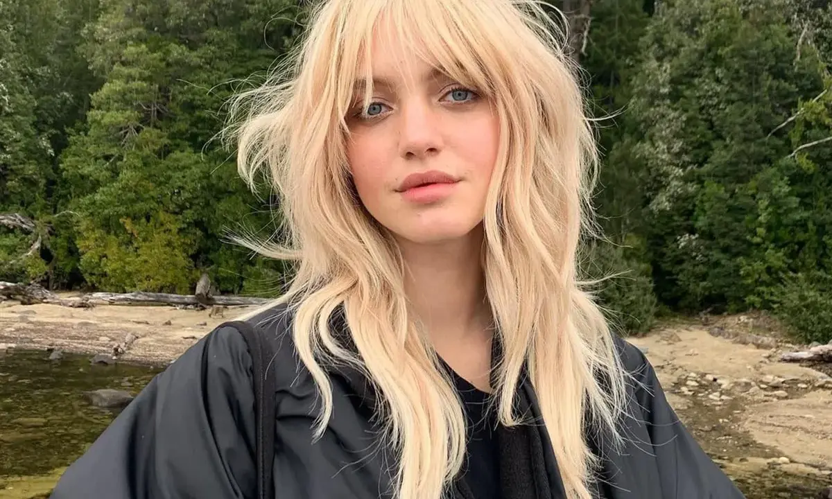Blonde Frau mit lässigem Wolf Cut stylen, blauen Augen und einem schwarzen Jackett vor einer Waldkulisse.