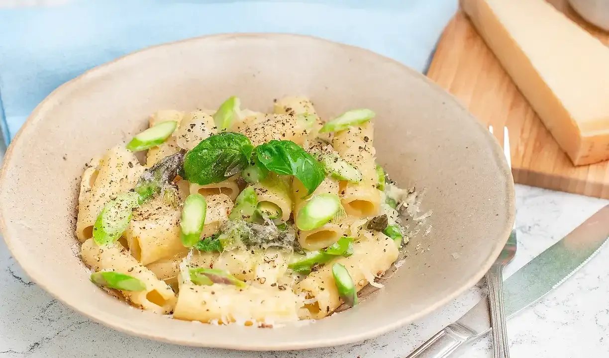 Creamy pasta with asparagus, basil, and grated parmesan cheese in a bowl, with a wedge of parmesan on a cutting board.