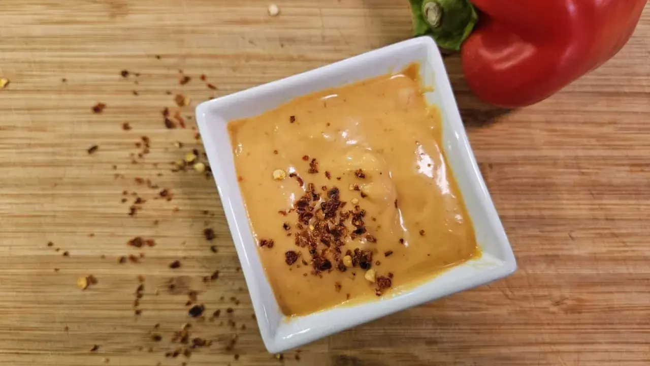 Cremige Chipotle-Sauce in einer weißen Schale, bestreut mit Chiliflocken. Daneben liegt eine rote Paprika auf einem Holzbrett.