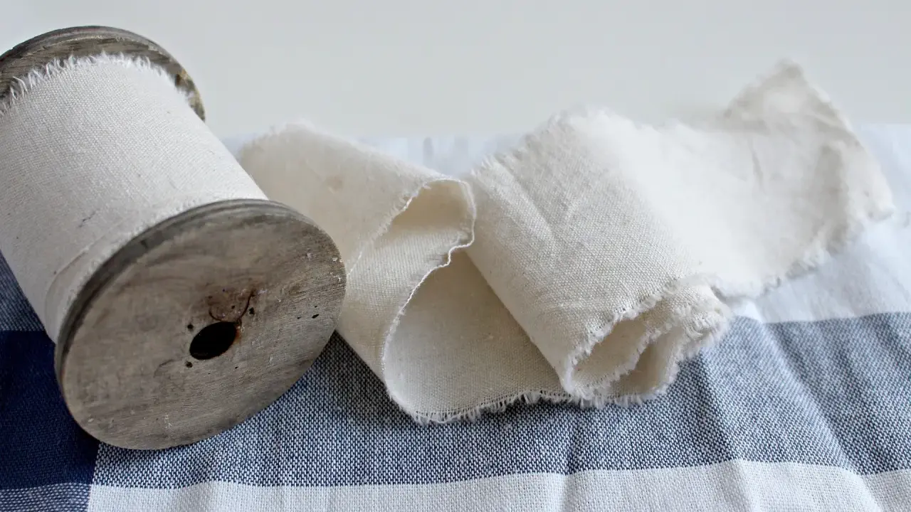 Un vecchio rocchetto di legno con tessuto di cotone pettinato arrotolato. Il tessuto è grezzo e sfrangiato, adagiato su una tovaglia a quadri blu e bianchi.