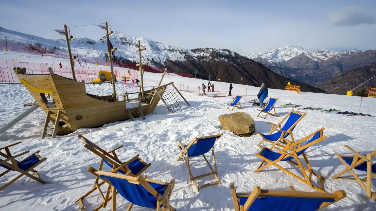 Alpe di Mera: un galeone di legno e sdraio blu attendono i visitatori sulla neve, con montagne innevate sullo sfondo.