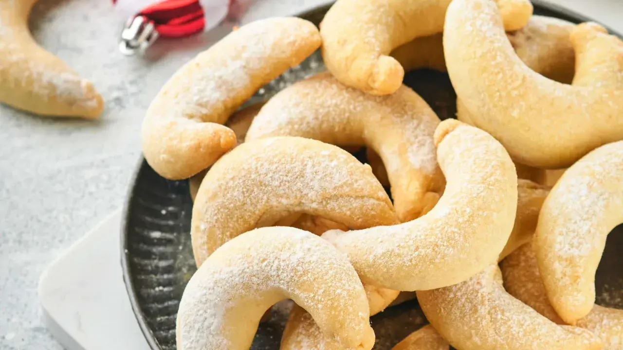 Ein Haufen goldbrauner Vanillekipferl, bestäubt mit Puderzucker, liegt auf einem dunklen Teller. Im Hintergrund sind weihnachtliche Zuckerstangen zu sehen.