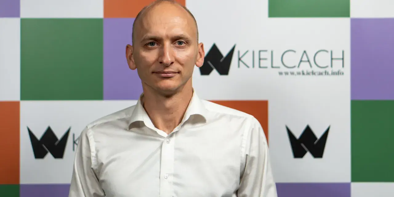 Łysy mężczyzna w białej koszuli na tle kolorowej ściany z logo "wKielcach".