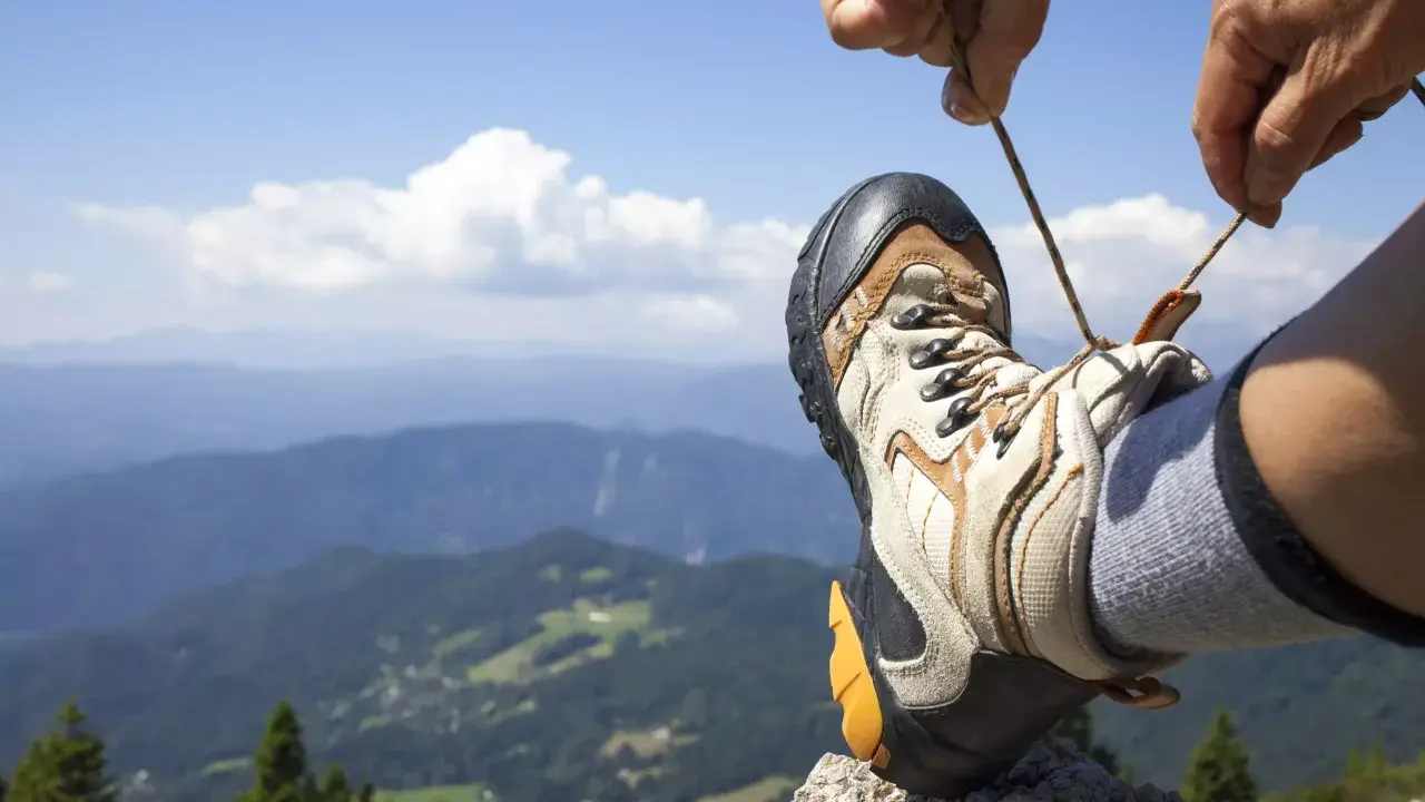 Ręka zawiązuje sznurówki buta trekkingowego na tle górskiego krajobrazu. Właściwe wiązanie butów to klucz do komfortowej wędrówki.