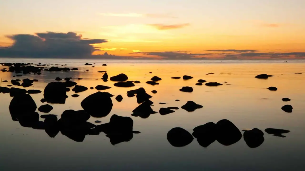 Solnedgång över havet vid en stenig strand på ar Gotland. Mörka stenar speglas i det lugna vattnet under en himmel i orange och gult.