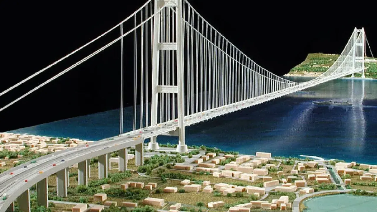 Maqueta de un puente colgante con pilares y cables que forman **puentes triangulados**, cruzando un cuerpo de agua y conectando dos orillas con una ciudad en miniatura.
