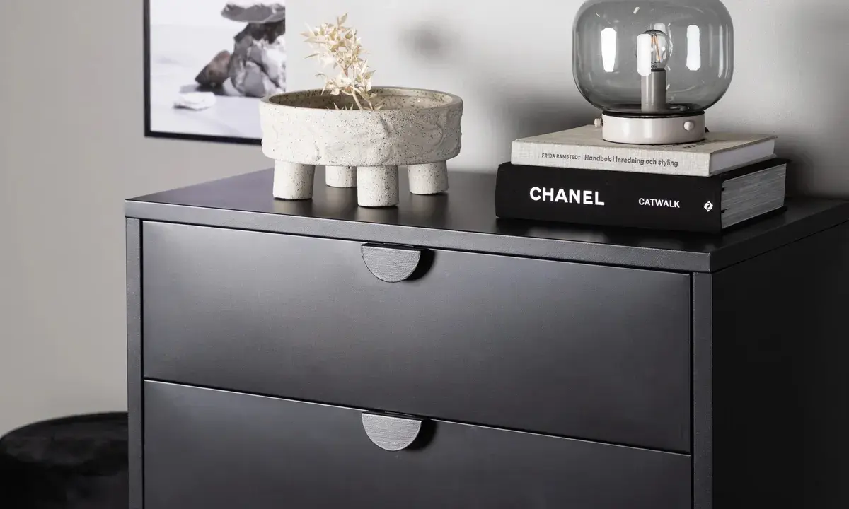 Svart sovrum med en mörk byrå. På byrån står en kruka med torkad växt, en lampa och böcker, bland annat "CHANEL CATWALK".