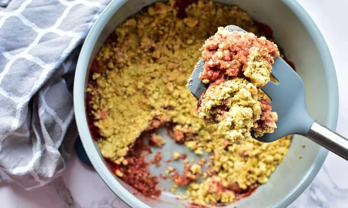 Ein köstlicher Erdbeer Rhabarber Crumble wird mit einem Löffel aus einer Auflaufform gehoben. Daneben liegt ein gemustertes Küchentuch.