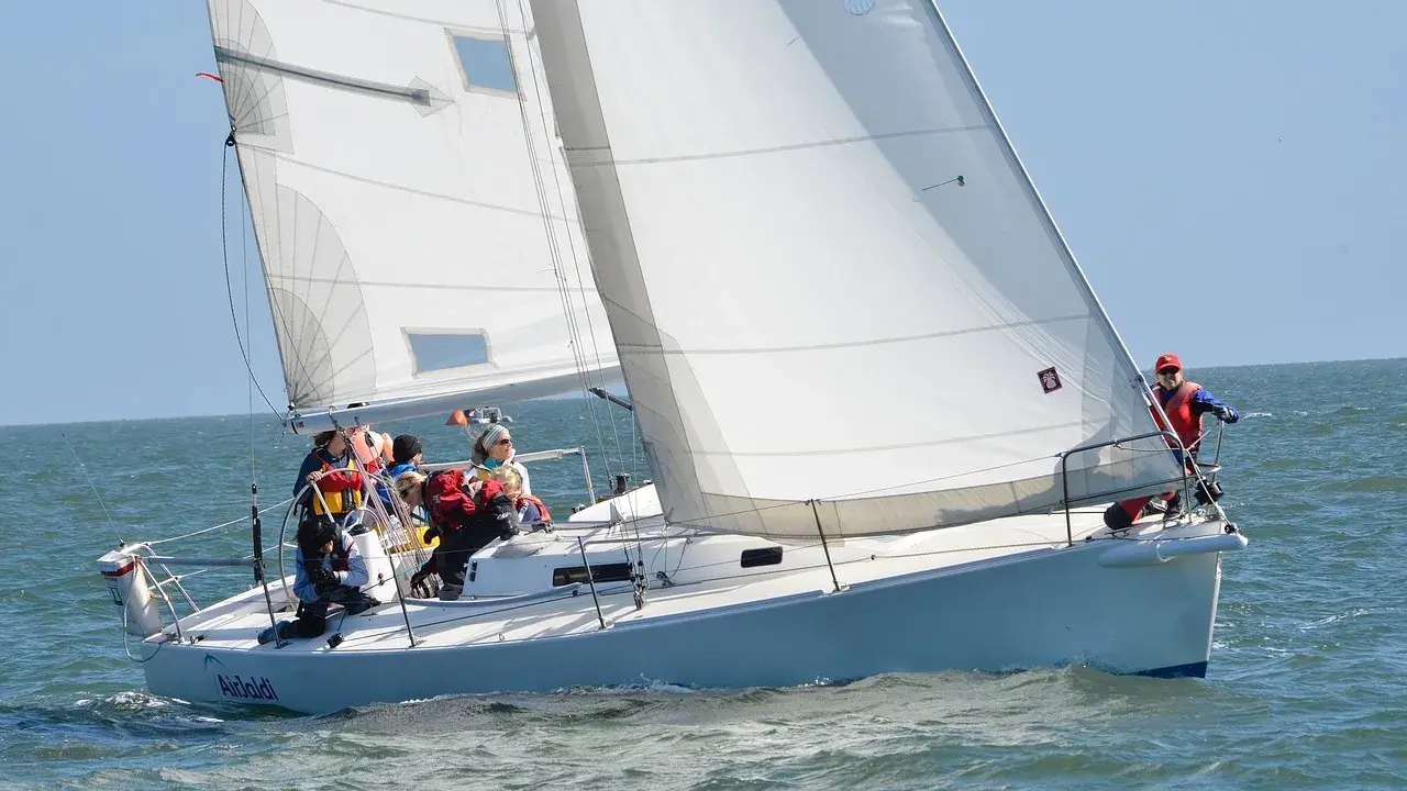 Un voilier blanc navigue sur une mer agitée, avec plusieurs personnes à bord. Le soleil brille, les voiles sont gonflées par le vent.