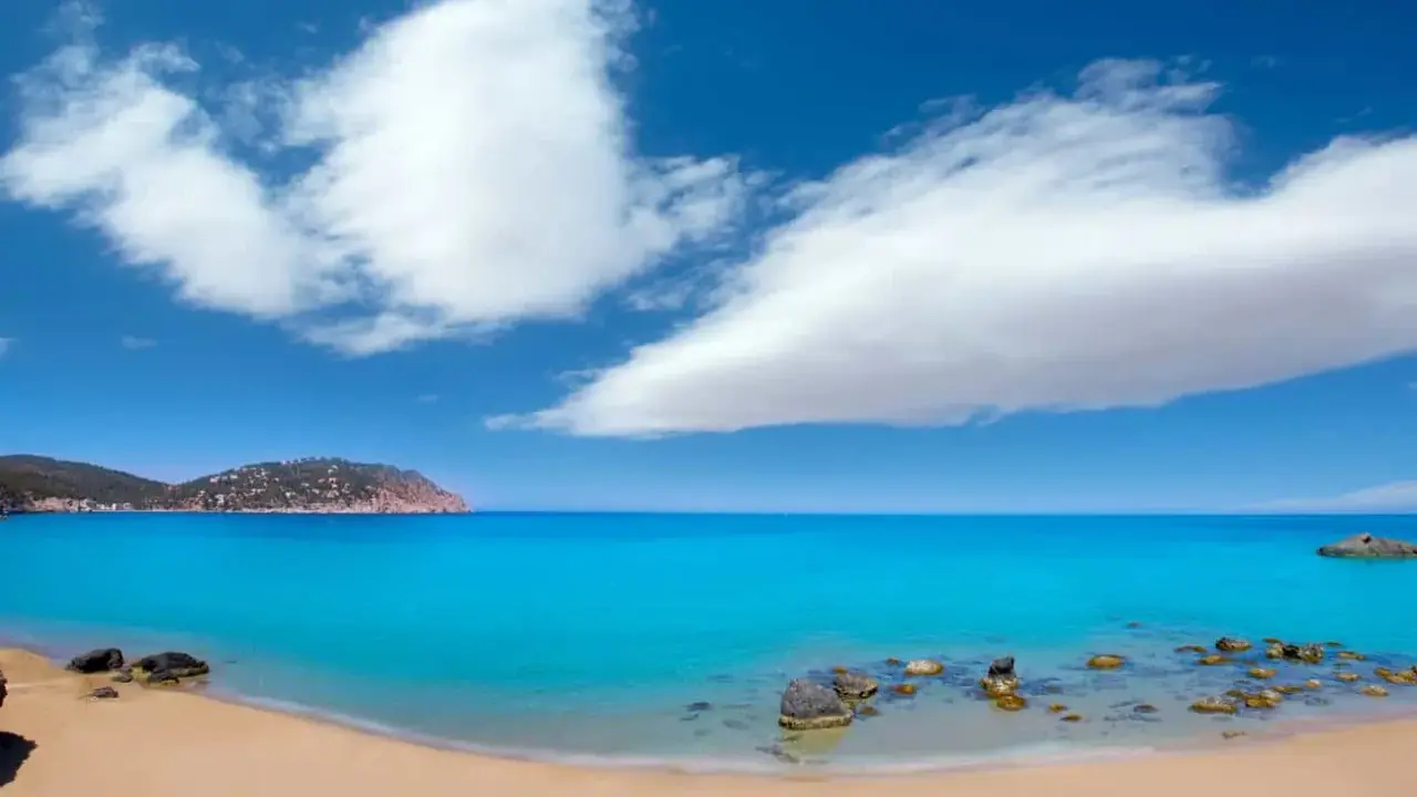 Traumhafter ibiza strand mit türkisblauem Wasser, goldenem Sand und einer felsigen Küste unter einem strahlend blauen Himmel mit weißen Wolken.