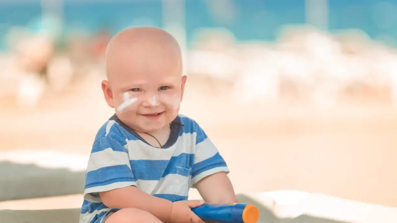 Un bébé souriant avec de la crème solaire sur le visage, tenant un tube de crème solaire. Idéal pour savoir crème solaire bébé à partir de quel âge.