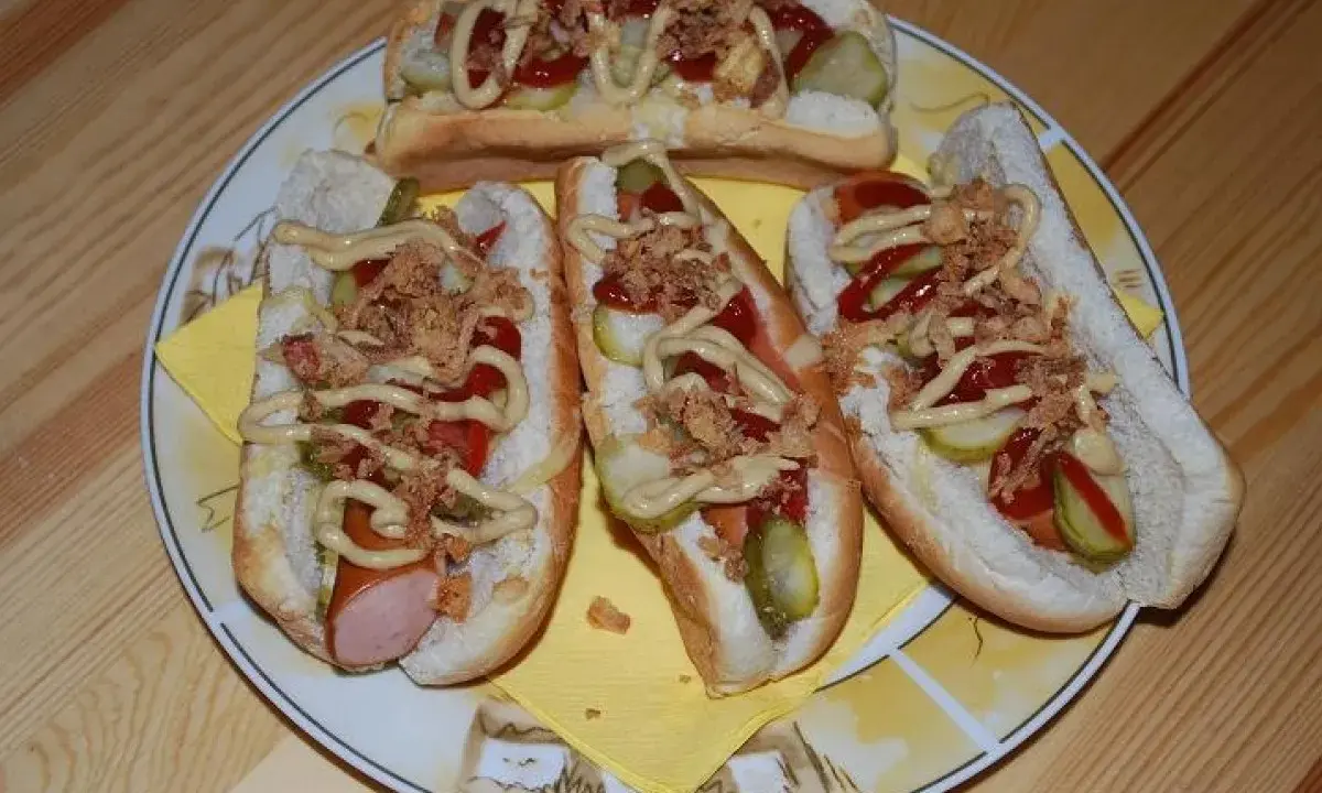 Cztery pyszne domowe hot dogi z kiełbaską, ogórkiem, ketchupem, musztardą i chrupiącą cebulką na talerzu.
