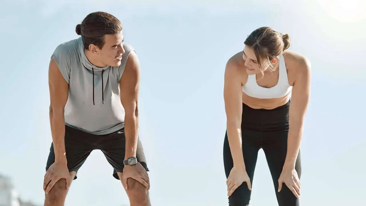 Zwei Läufer erholen sich nach einem anstrengenden 10 km Lauf. Sie schauen sich an, die Hände auf den Knien, erschöpft, aber zufrieden.
