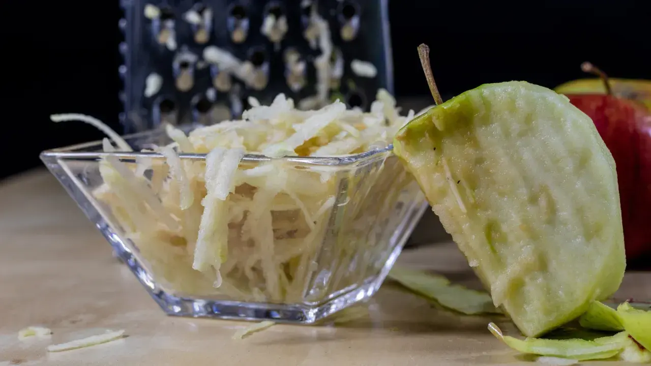 Frisch geriebener Apfel in einer Glasschale, daneben ein Apfelstück und eine Reibe.