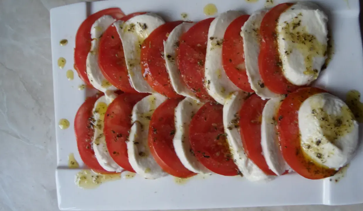 Mozzarella z pomidorami, ułożona naprzemiennie na białym talerzu, skropiona oliwą i ziołami.