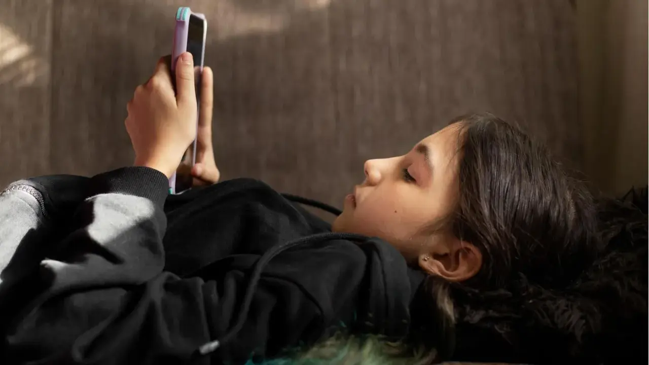 Ein Mädchen liegt auf einer Couch und schaut auf ihr Smartphone. Sie ist in ein schwarzes Kapuzensweatshirt gekleidet. Dies spiegelt aktuelle Trends der Jugend wider.