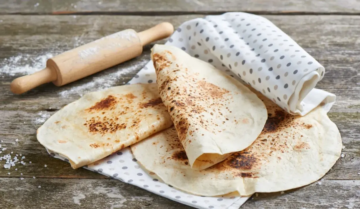 Świeżo usmażona tortilla z patelni, lekko przypieczona, leży na lnianej ściereczce obok wałka.