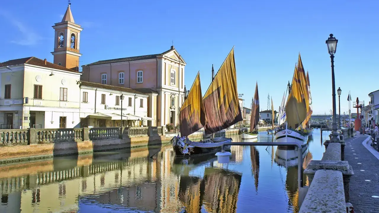 Barche a vela tradizionali ormeggiate sul canale di Cesenatico mare, con edifici storici e riflessi sull'acqua.