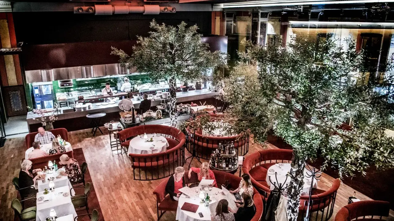 Elegant restaurang med runda, röda bås och träd. Köket syns i bakgrunden. Många gäster njuter av sin måltid. L'avventura recensioner verkar positiva.