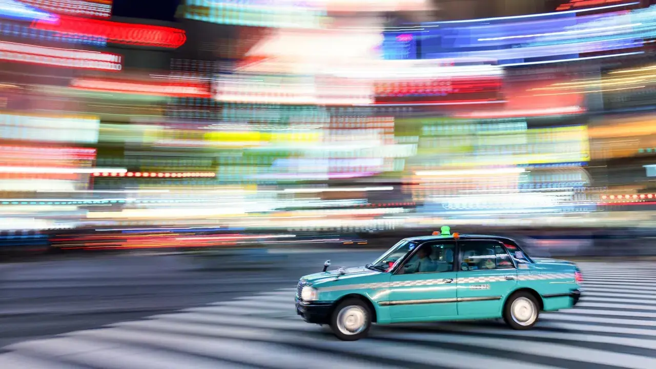 Un taxi azzurro sfreccia su strisce pedonali, con luci sfocate di città che creano un effetto dinamico, perfetto per imparare come fare foto in movimento.