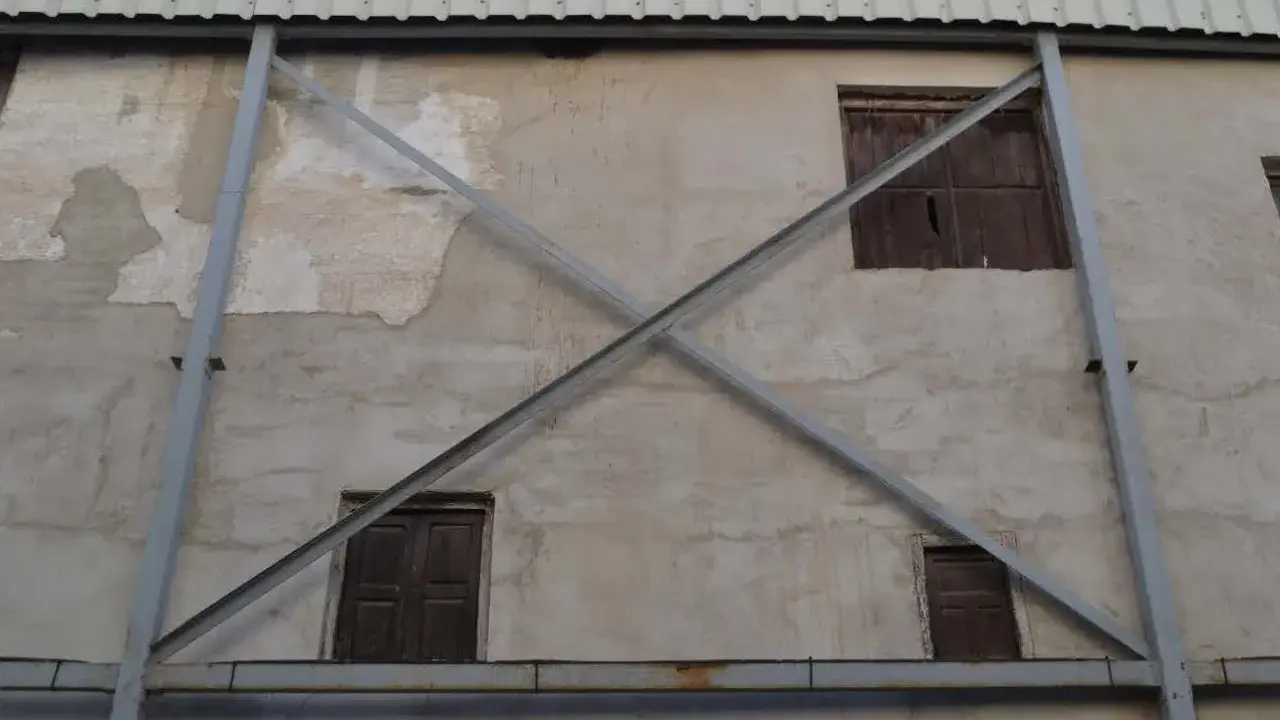 Estructura metálica en forma de X que refuerza una pared de concreto con ventanas de madera. La riostra, en este caso, es un elemento de soporte crucial.