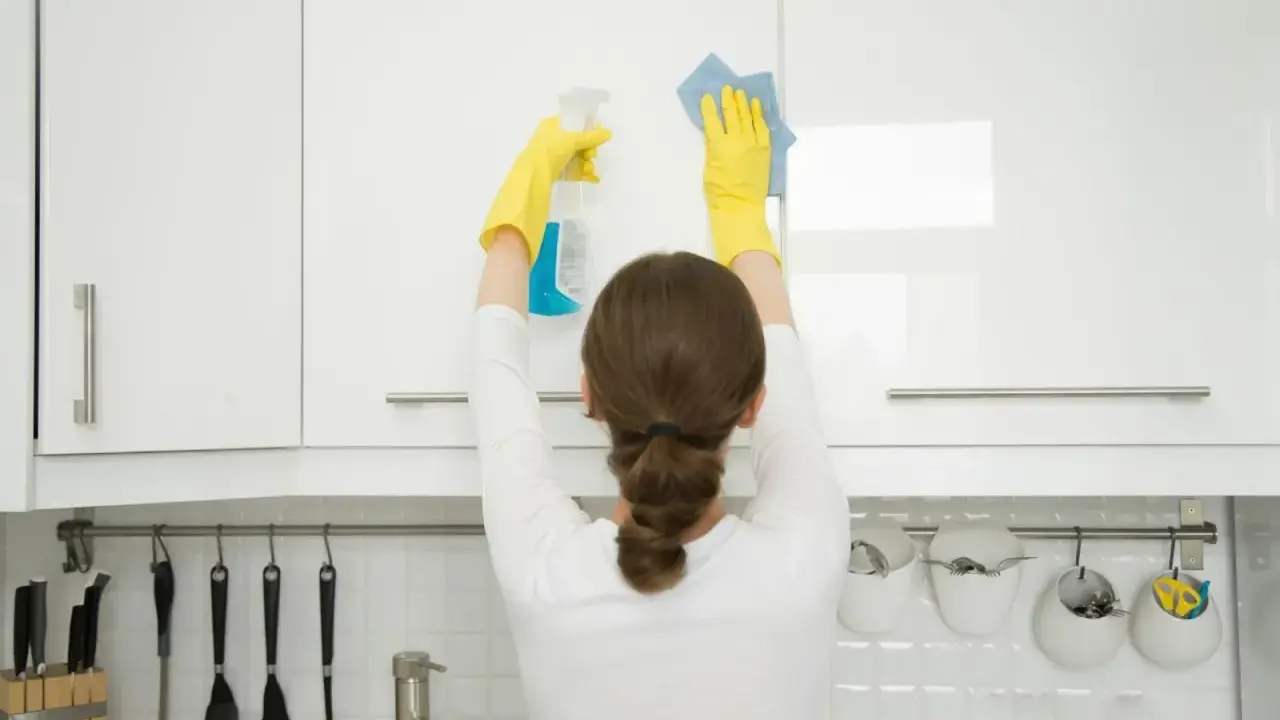 Mujer con guantes amarillos realiza limpieza armarios cocina, rociando y limpiando con paño azul.