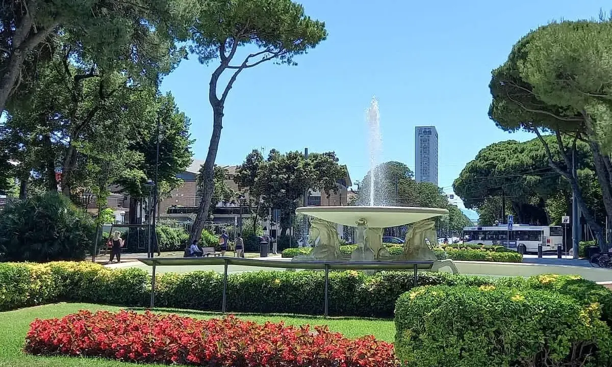 Fontana z wodą w **Parco Fellini Rimini**, otoczona zielenią i kwiatami. W tle widać autobus i wieżowiec.