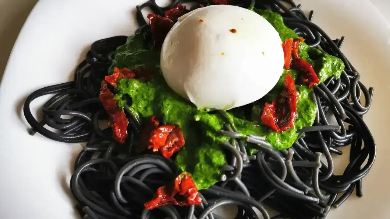 Czarny makaron z zielonym pesto, suszonymi pomidorami i kulką mozzarelli.