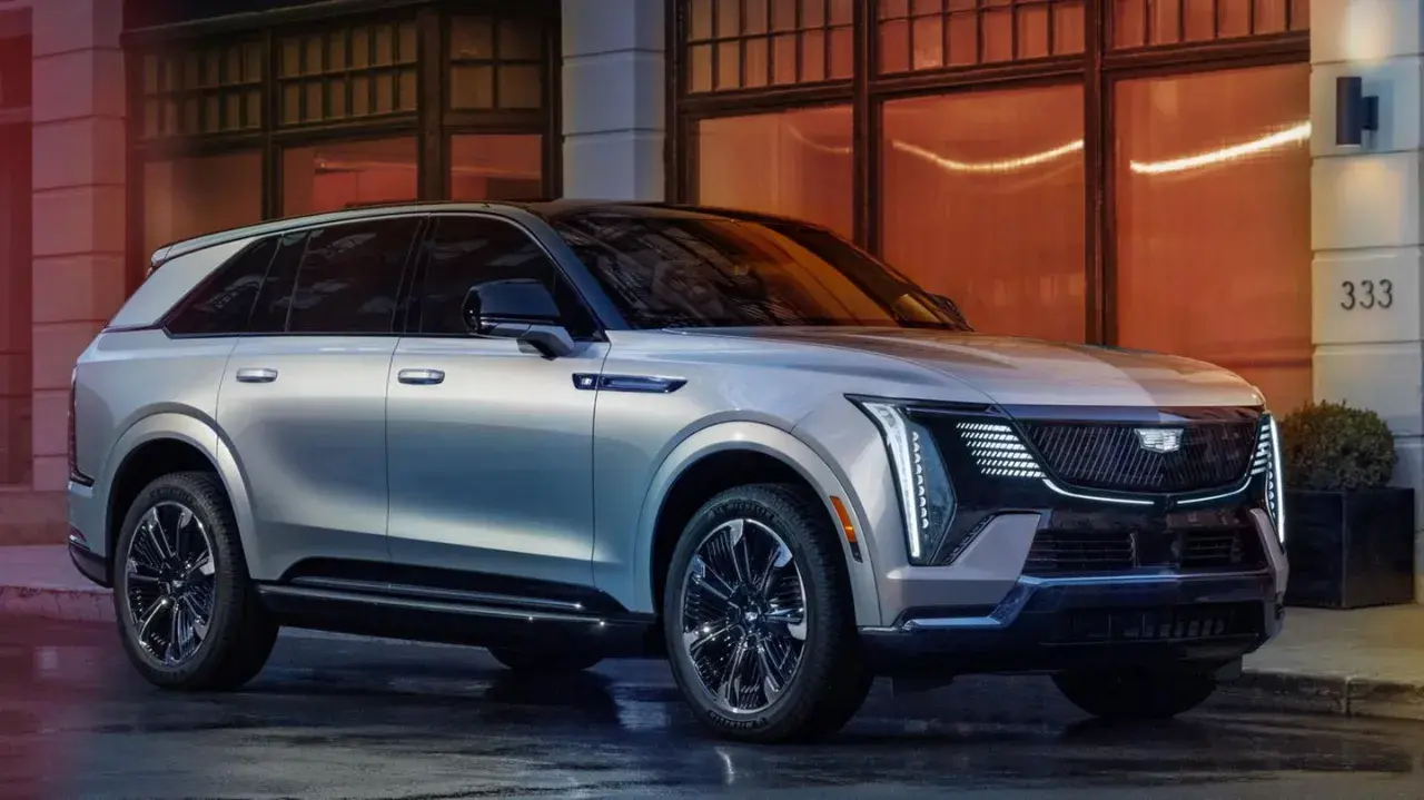 Eleganter silberner Cadillac Escalade IQ parkt vor einem Gebäude mit orangefarbenen Fenstern.