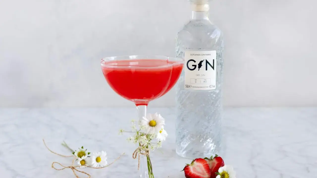 En röd drink i ett coupeglas, bredvid en flaska gin från Gotlands Ginfabrik. Jordgubbar och små blommor dekorerar bordet.