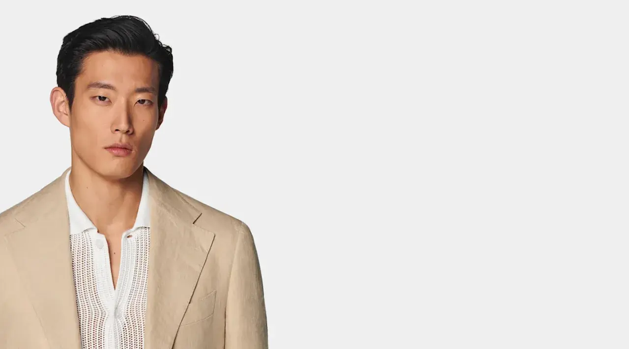 A man models a tan linen blazer over a white textured polo shirt, perfect for a smart casual dress code for a wedding.