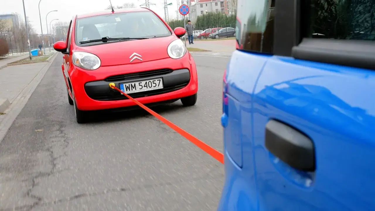 Czerwony Citroen holowany przez niebieski samochód. Pomarańczowa linka holownicza jest napięta, sugerując ruch i prędkość.